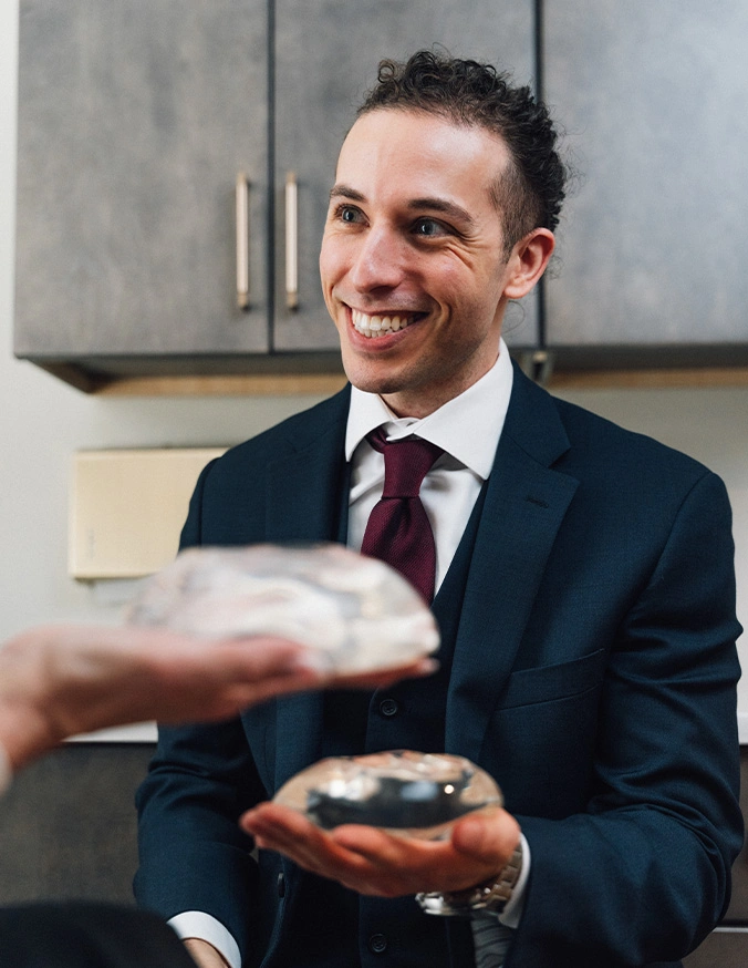 Doctor smiling while holding a breast implant and another hand is holding a implant out in front of him. - Breast implant removal in Wayne, PA