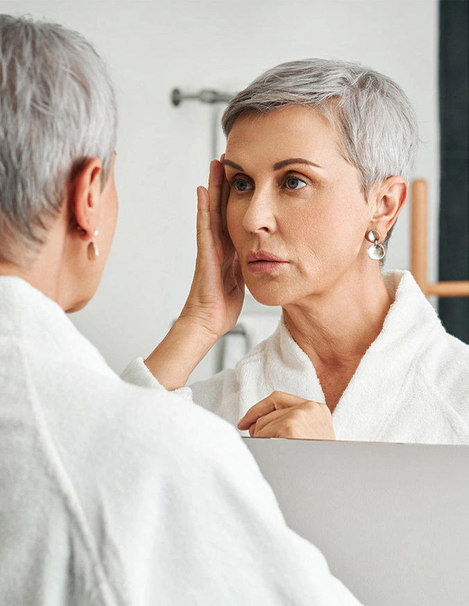 Woman with short gray hair examining her face in bathroom mirror while wearing white robe - C02 laser in Wayne, PA