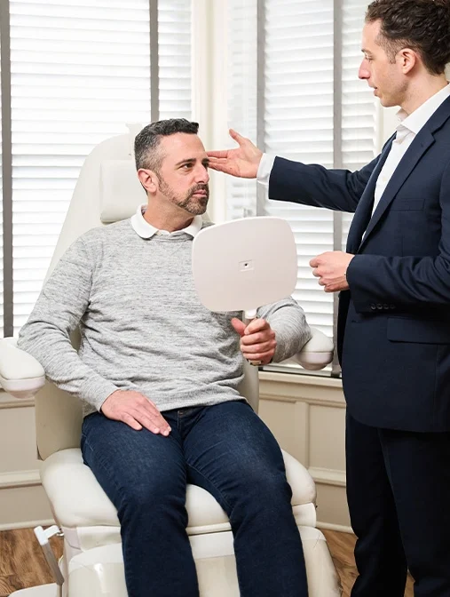 Doctor Marc Govert looking at male patient in a medical chair as the patient holds up a mirror. - Eyebrow restoration in Wayne, PA