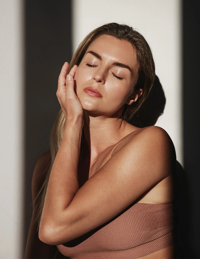 Woman with glowing skin and natural makeup touching face in soft sunlight, wearing neutral tone top - IPL in Wayne, PA