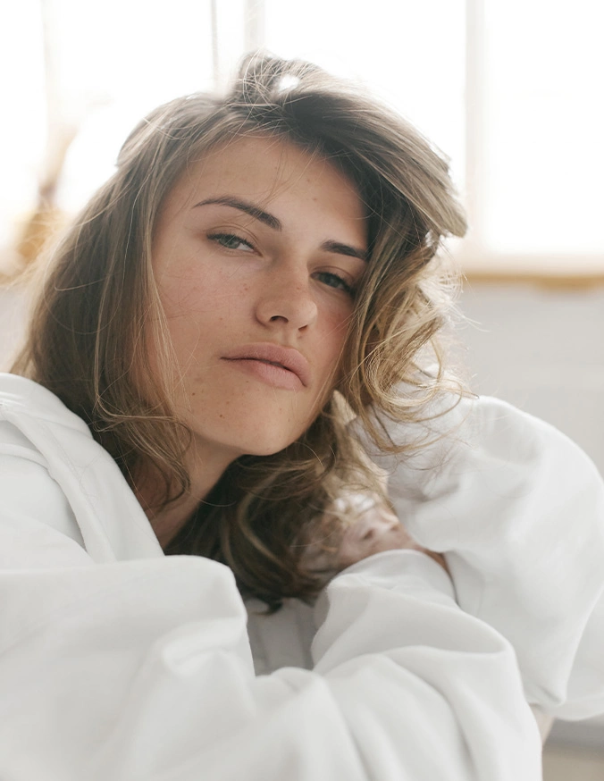 Woman with wavy brown hair in white bathrobe looking contemplative in natural lighting - Lip fillers in Wayne, PA