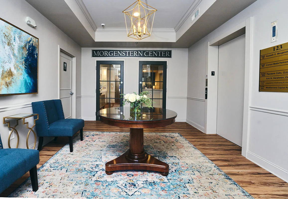 Modern entryway with blue velvet chair, round wooden table on colorful rug, and elegant chandelier lighting