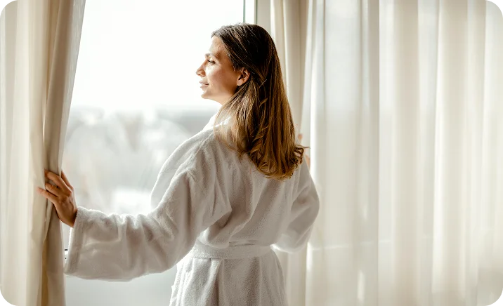 Woman in white bathrobe opening curtains by window in bright hotel room or spa - out of town in Wayne, PA