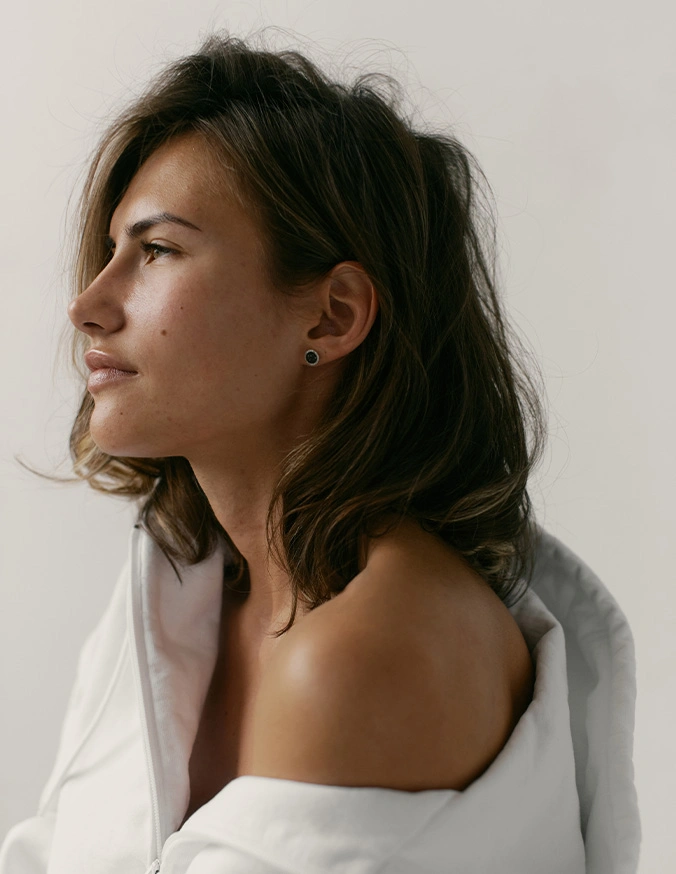 Woman in profile view with natural makeup and tousled hair wearing white off-shoulder top against neutral background - Rhinoplasty in Wayne, PA